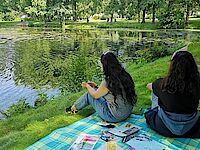 2 jugendliche Mädchen auf einer bunten Picknickdecke im Park schauen über den Teich